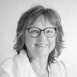 Smiling woman with short gray hair and black-rimmed glasses, wearing a light blouse.
