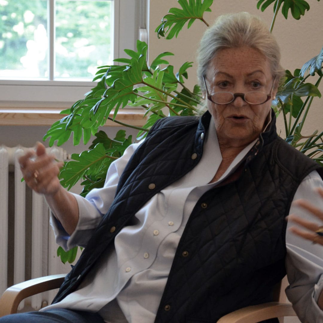 Older woman with reading glasses perched low on her nose, gesturing while seated indoors with a large leafy plant behind her.