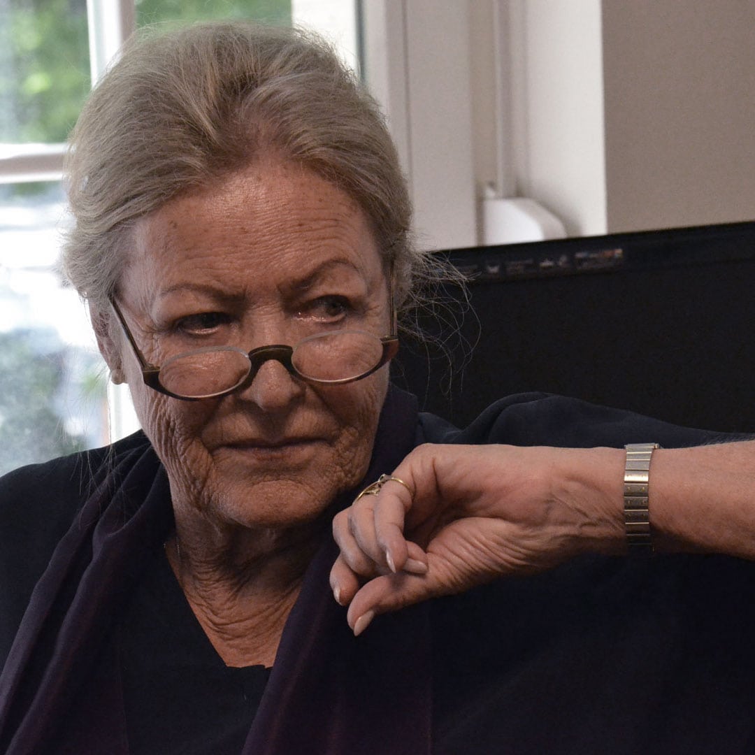 Close-up of an elderly woman with gray hair and glasses, looking at her wristwatch.