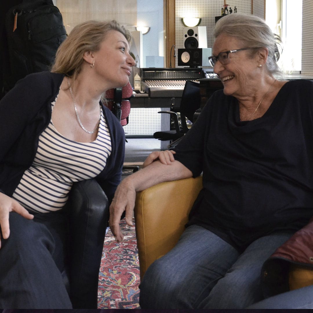 Two women smile as they chat in a recording studio lounge with audio gear nearby.