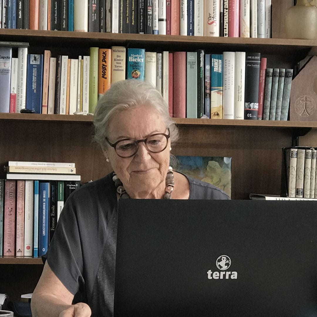 Older woman with glasses sits at a desk using a black Terra laptop in a library/study filled with books behind her.