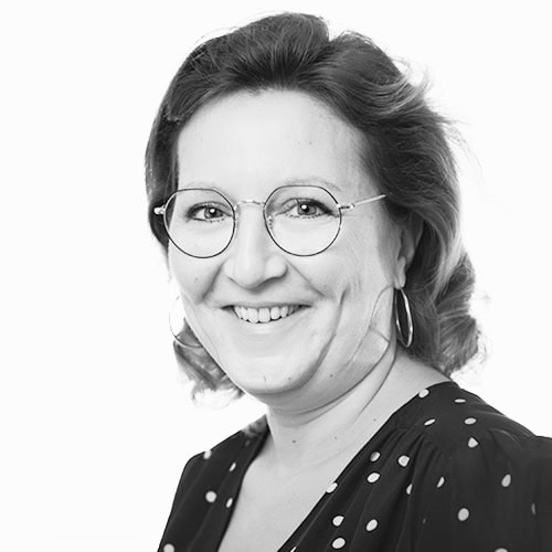 Smiling woman with round glasses and hoop earrings, wearing a polka-dot blouse, photographed in grayscale.