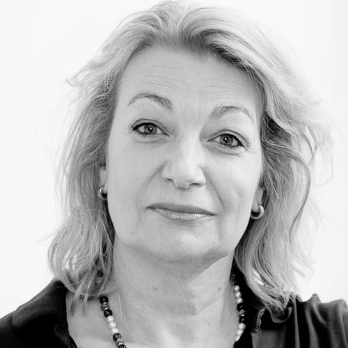 Black-and-white portrait of a middle-aged woman with shoulder-length light hair and a soft smile, wearing a pearl necklace and small hoop earrings.