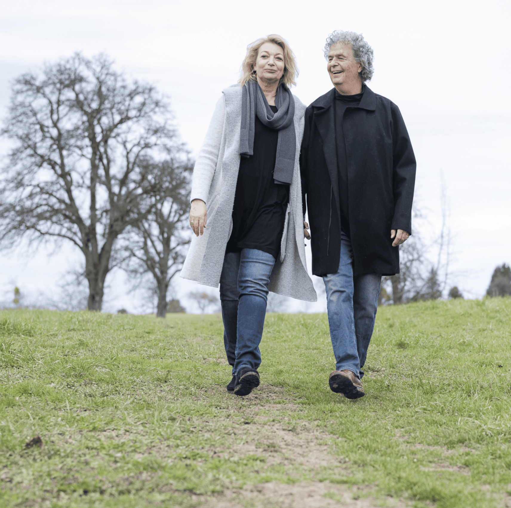 A man and a woman walking together on a grassy trail, smiling, on a cool day with bare trees in the background.